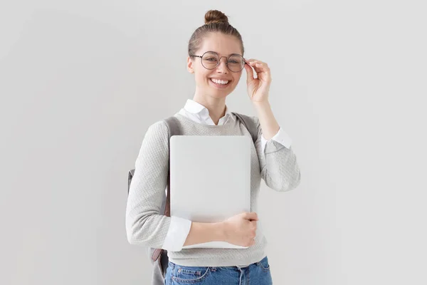 Innenaufnahme eines Studentenmädchens, das isoliert auf grauem Hintergrund mit geschlossenem Laptop in der Hand steht, die Brille mit offenem positivem Lächeln gerade setzt und Begeisterung und Initiative ausdrückt. — Stockfoto