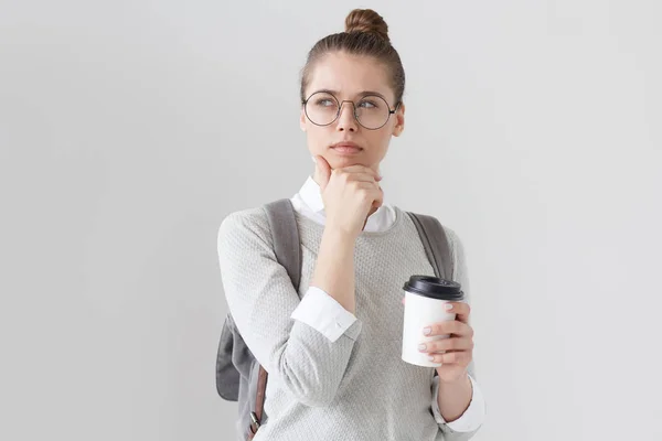 Horizontales Foto einer jungen, gut aussehenden Europäerin mit Rucksack und schwarz umrandeter Brille auf grauem Hintergrund, die misstrauisch, unsicher und voller Zweifel an der aktuellen Situation wirkt. — Stockfoto