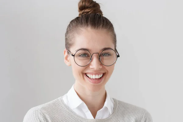 Foto interior de atraente jovem morena europeia com olhos verdes isolados em fundo cinza, cabelo amarrado em coque, com óculos grandes, rindo, sorrindo abertamente e expressando respeito ao interlocutor . — Fotografia de Stock