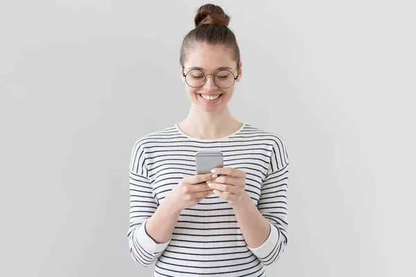 Närbild foto av nonchalant klädd europeisk flicka står isolerad på grå bakgrund tittar uppmärksamt på skärmen av mobiltelefon, bläddra webbsidor och ler fint medan chatta med vänner. — Stockfoto