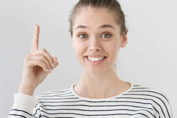 Närbild skott av trevligt leende unga kaukasiska flicka isolerad på grå bakgrund pekande finger upp till uppmärksamma viktig information eller ny idé eller erbjudande, letar efter engagerade och ivriga att dela. — Stockfoto