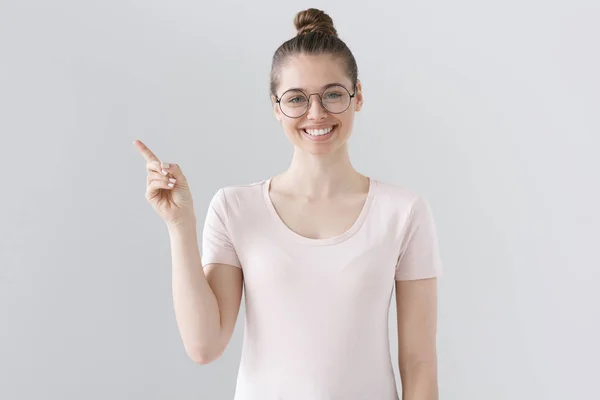 Retrato de una bonita mujer europea en camiseta rosa aislada sobre fondo gris, con gafas redondas, llamando la atención sobre algo a la izquierda como si lo recomendara en base a una experiencia personal positiva . — Foto de Stock