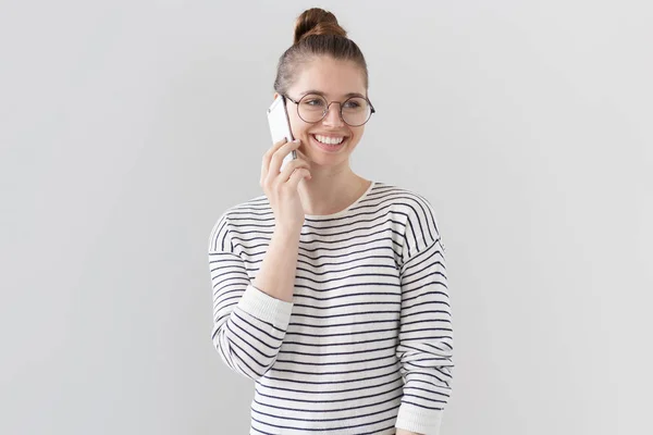Binnen foto van mooie jonge Europese vrouwelijke permanent geïsoleerd tegen de grijze achtergrond in grote ronde bril, zijwaarts kijken en praten op mobiele telefoon met blije uitdrukking op het gezicht en vrolijke glimlach. — Stockfoto
