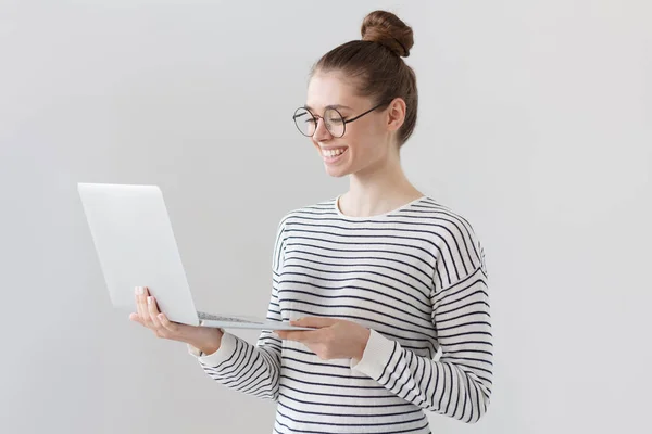 Shot of beautiful young girl standing isolated against gray background wearing big round glasses, standing and looking at screen of laptop that she holds, smiling happily at positive news from web. — Stock Photo, Image