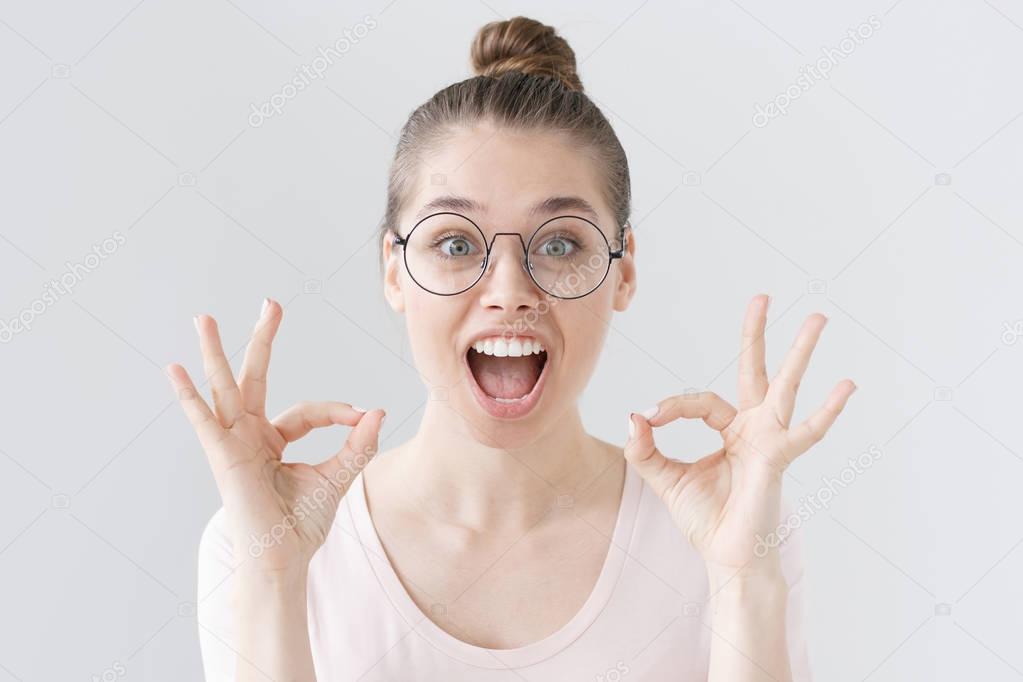 Photo of young European woman isolated on grey background in thin-framed eyeglasses with emotional expression of surprise and joy, her mouth and eyes wide open as she sees unbelievable things.