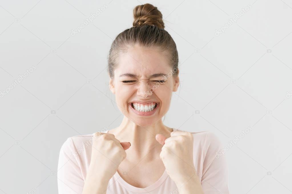 Closeup of emotional European girl isolated on gray background showing white teeth while screaming with joy and victorious expression, holding hands in gesture of winner, looking extremely happy.