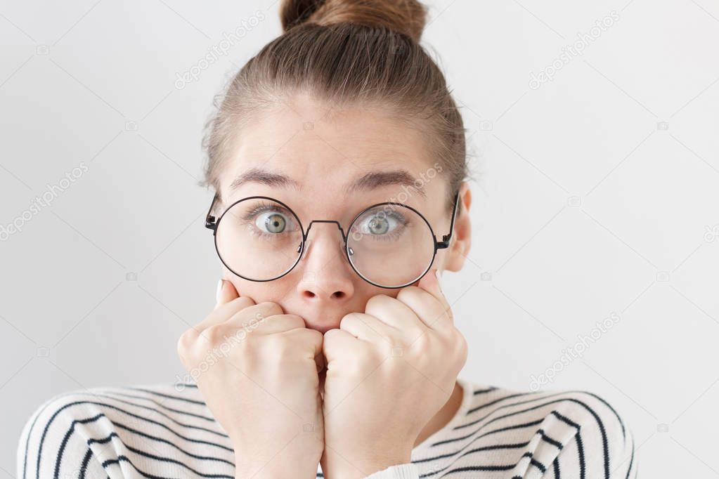 Shot of frightened female in round black-rimmed eyeglasses isolated on grey background astonished with unusual and important news, touching her face with fingers, hiding mouth open wide in surprise.