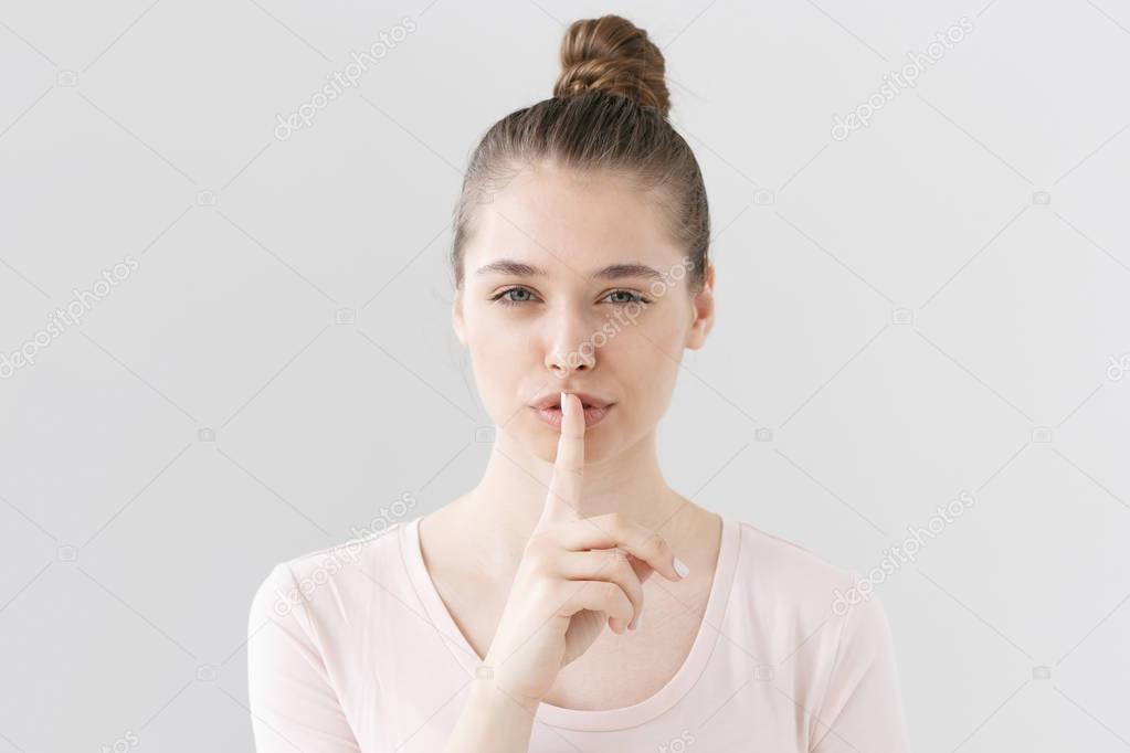Indoor portrait of beautiful European female isolated on gray background pressing forefinger to lips to make interlocutor silent, smiling intricately with secrecy as if promising something exciting.
