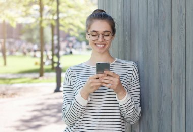 Çizgili üst dikkatle yazarak, akıllı telefon, sergilenen memnun gülümseme ile net dostu neşeli iletişime devam etmek isteyen bakıyorum giymiş yakışıklı beyaz kız portre fotoğrafı.