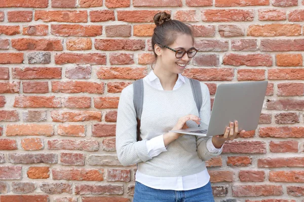 Closeup photo of young European female standing against red brick background smiling happily while holding laptop in hands, using touchpad to browse in web and reply to messages from friends. — Stock Photo, Image