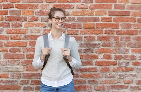 Foto ao ar livre de jovem bela senhora de pé contra a parede de tijolos vermelhos com mochila cinza, vestindo roupas neutras casuais, jeans e óculos de moda com chifres, olhando para a direita como se estivesse esperando . — Fotografia de Stock