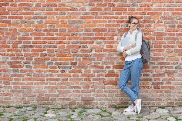 Full-height portrait of young beautiful woman leaning to red brick wall holding laptop in hands and turning rightwards as if awaiting for friend to come, looking happy and relaxed during her walk.