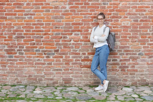Full-height portrait of young European woman wearing eyeglasses and casual clothes, leaning to red brick wall with her backpack on, smiling confidently at camera willing to pose with arms crossed.