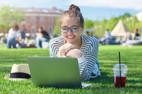 Closeup portrait of young beautiful female looking at laptop display with excitement and gesture of impatience and involvement while lying on grass in park and drinking berry juice, enjoying sun. — Stock Photo, Image