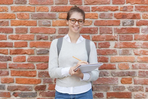 Imagem de close-up da jovem mulher europeia em pé contra o fundo de tijolo vermelho vestindo camisa branca e pulôver cinza, segurando notebook e lápis para esboçar com sorriso aberto cheio de inspiração . — Fotografia de Stock