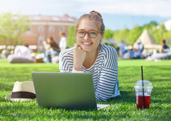 Summer closeup of smiling good-looking positive girl in trendy eyeglasses and striped top, spending free time on green lawn with open laptop, straw hat lying beside her and plastic cup of fresh juice. — Stock Photo, Image