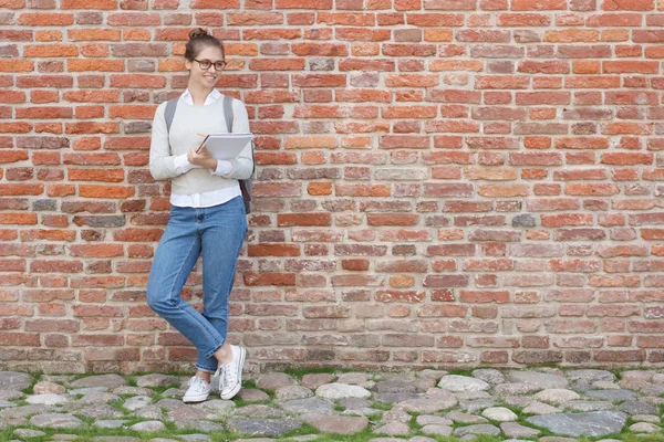 Full-height photo of young beautiful Caucasian female pictured on the left with red brick behind, showing relaxed smile while looking at object she is going to draw with pencil in her notebook.
