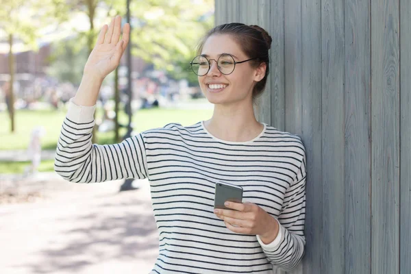 Outdoor image of young happy European woman greeting mate she sees in street with one hand and holding cellphone in other, wearing big thin-rimmed trendy eyeglasses with friendly positive smile. — Stock Photo, Image