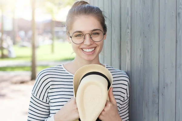 Closeup photo of young smiling European female leaning to gray wooden wall with shoulder, holding sun hat in hands while listening to funny jokes from friend and reacting with happy laughter. — Stock Photo, Image