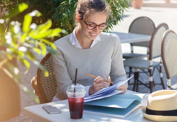 Closeup photo of young beautiful European woman spending time in outdoor cafe one sunny summer day looking through important papers on table and writing with pencil with happy positive smile — Stock Photo, Image