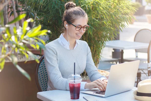 Venkovní portrét mladé usměvavé evropské ženy u stolu café, obklopený zelení, její laptop otevřené, poslechu hudby přes sluchátka, pití šťávy a užívání si letního času venku — Stock fotografie