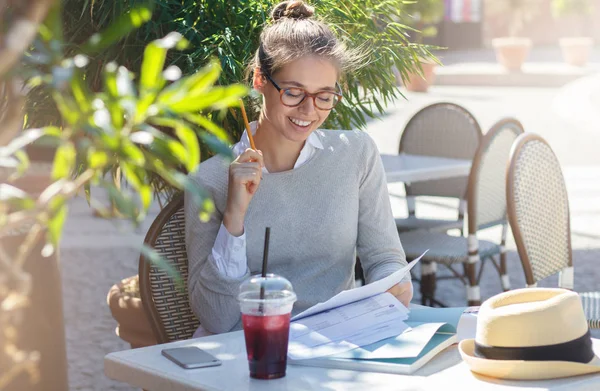 Outdoor portrait of young good-looking Caucasian female in glasses sitting at cafe table outdoors and filling in forms on papers with pencils feeling happy about both working and having nice rest — Stock Photo, Image