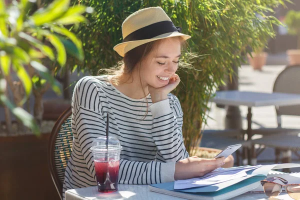 Closeup photo of shy beautiful girl looking at display of her cellphone while exchanging content and messages in web as she is spending leisure time in outdoor cafe, drinking fresh berry juice — Stock Photo, Image