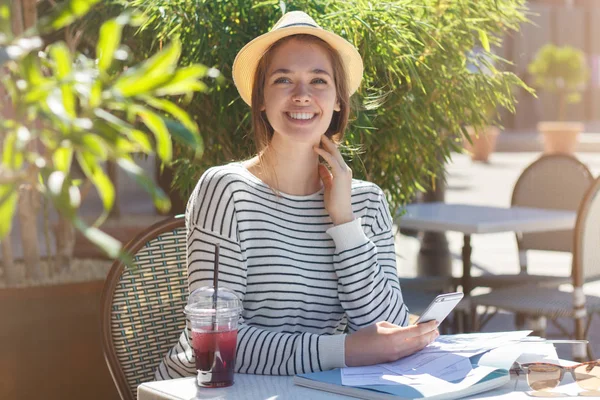 Closeup image of young beautiful female wearing hat sitting at table in street cafe in summer, holding phone, looking to what is happening in street with anticipating tender smile as if seeing friend — Stock Photo, Image