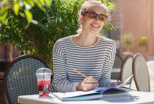 Outdoor closeup of good-looking European girl in trendy sunglasses laughing and looking excited and happy when reading papers and printed materials lying on table in street cafe, writing with pencil — Stock Photo, Image