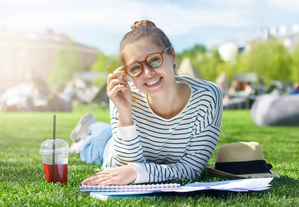 Bright summer closeup of smiling European student girl in trendy eyeglasses looking straight at camera while preparing for classes in city park on green lawn, drinking berry juice and enjoying time — Stock Photo, Image