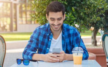 Genç yakışıklı Avrupa erkek Tshirt ve taze meyve suyu içme ve mutlu bir şekilde gülümseyerek arkadaş ile smartphone, aracılığıyla ileti açık kafe dinlenme sahip mavi damalı gömlek closeup portresi