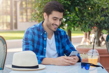 Boş zaman zaman mutlu kahkaha ile cep telefonu ekranına bakarak, dinlenme ve kendini zevk sokak Cafe masada rahat giyen genç olumlu beyaz adam açık closeup elbise