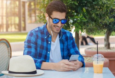 Taze meyve suyu içme ve sokulgan yolu ile cellphone kulaklık ile temas kalmayı açık sokak Cafe geçiriyor yatay güneşli yaz öğleden sonra zevk mutlu yakışıklı çocuk portresi