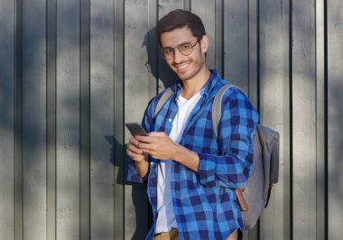 Genç olumlu Avrupa adam sırt çantası, rahat kıyafetler ve konuşma zevk veya smartphone üzerinden web tarama gözlük gri ahşap çit arka plan ayakta açık closeup