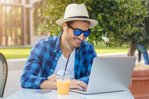 Foto de primer plano del joven guapo hombre caucásico mirando la pantalla de su computadora portátil y escuchando la voz a través de los auriculares con emoción mientras come en la cafetería al aire libre, sonriendo felizmente —  Fotos de Stock