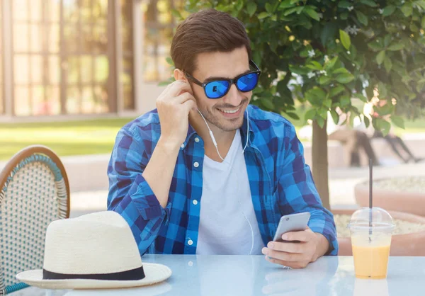 Imagen al aire libre del joven hombre caucásico sonriente que se comunica con su novia a través de auriculares de teléfono inteligente o viendo el contenido web mientras está sentado en la mesa de la cafetería de la calle y disfruta del tiempo libre en el día soleado —  Fotos de Stock