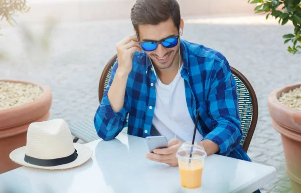 Крупный план молодого красивого белого мужчины в солнечных очках и клетчатой рубашке, проводящего свободное время за столом уличного кафе, смотрящего на экран мобильного телефона со счастливой улыбкой, расслабляющего — стоковое фото