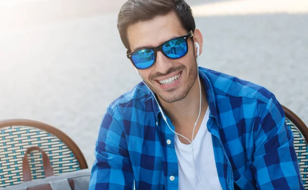 Horizontal picture of young handsome guy sitting in outdoor street cafe pictured with earphones on while talking to friend via cellphone laughing happily at jokes, enjoying relaxed and cozy atmosphere — Stock Photo, Image