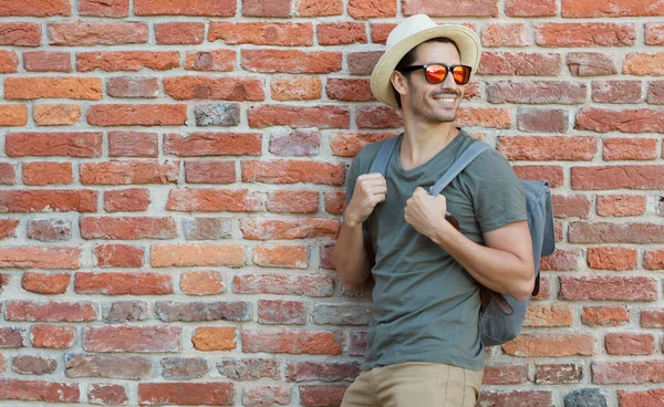 Outdoor image of positive European male standing in casual clothes with red brick wall behind, looking rightwards and holding backpack straps while discovering city centre and seeking new impressions — Stock Photo, Image