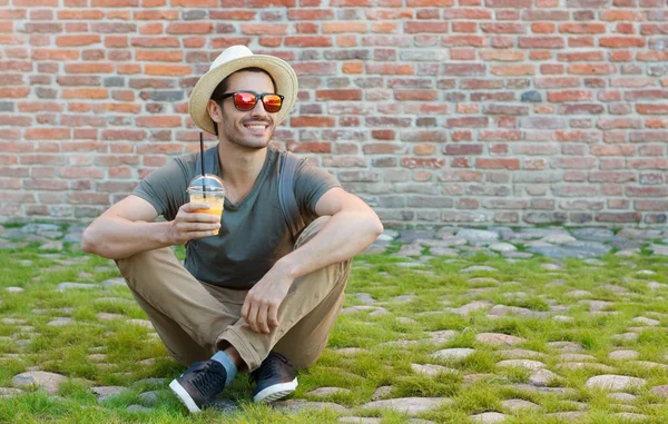 Horizontal closeup of young Caucasian man pictured on left side sitting on grass and stones with red brick background behind drinking fresh juice from plastic cup and watching street with interest — Stock Photo, Image