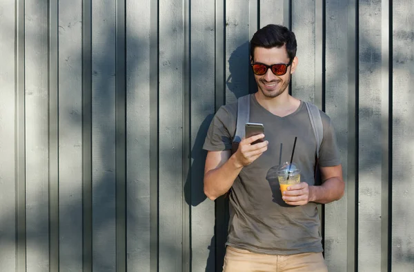 Horizontal outdoor image of young optimistic European man pictured with grey wooden fence behind, looking at screen of phone, browsing and looking through web content while staying in countryside — Stock Photo, Image