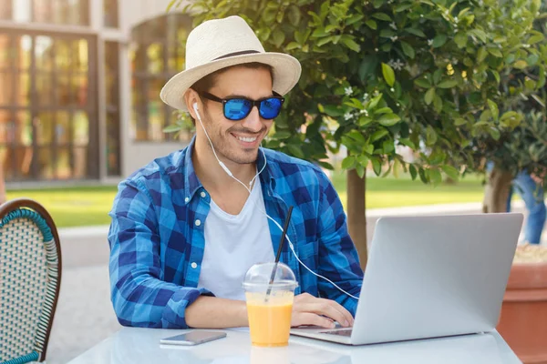 Retrato al aire libre de un joven europeo sonriente sentado en la mesa de la cafetería rodeado de vegetación, su portátil abierto, escuchando música a través de auriculares, bebiendo jugo y disfrutando de la hora de verano al aire libre —  Fotos de Stock