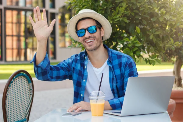 İyimser beyaz açık yatay closeup resimde sokak café'de tablo güneş şapka ve dostu onun tanıdık selamlıyorum ve ortak haberleri paylaşmak için gördüğü için sallayarak güneş gözlüğü takan adam