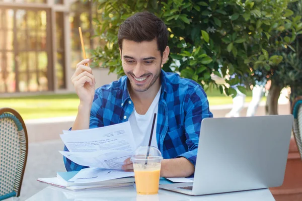 Outdoor portrait of young handsome Caucasian male sitting at cafe table outdoors and filling in forms on papers with pencils feeling happy about both working and having nice rest on sunny summer day