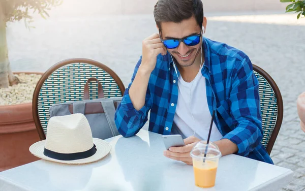 Imagen de primer plano del hombre europeo de moda en camisa casual azul sonriendo ampliamente mientras mira a la pantalla, cabeza doblada a teléfono inteligente, sentado en la mesa en la cafetería al aire libre en el día de verano soleado, disfrutando del tiempo libre —  Fotos de Stock