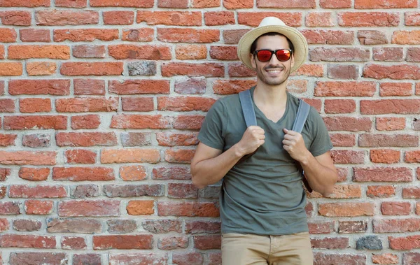 Outdoor portrait of young Caucasian man pictured with red brick wall in background looking through sunglasses with joyful smile, pulling straps of backpack forward as if ready to continue journey — Stock Photo, Image