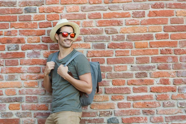 Horizontal image of young Caucasian man pictured on left side of red brick background watching what is happening in street walking in urban surrounding with backpack and hat on one summer day — Stock Photo, Image