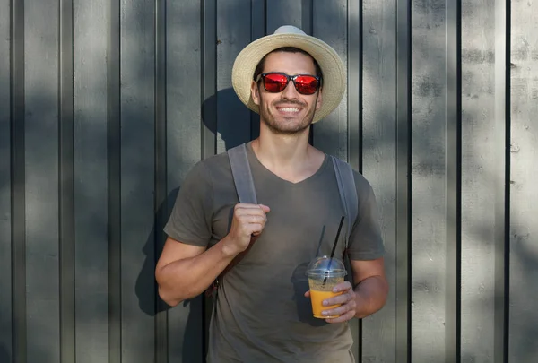 Outdoor picture of young European male standing in sun hat and sunglasses with gray wooden fence behind his back, holding strap of backpack and cup of drink, willing to continue walking around — Stock Photo, Image