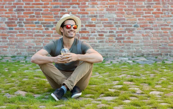 Outdoor portrait of positive European male having rest on pebbles and grass beside red brick background with plastic cup of fresh drink enjoying sunny summer day and spending time around town — Stock Photo, Image