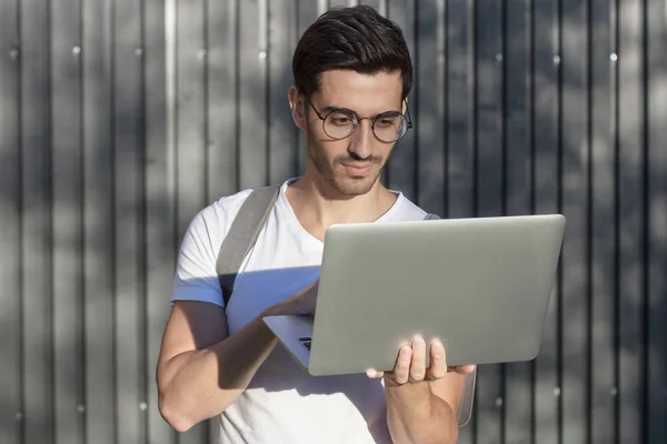 Retrato horizontal de un guapo hombre europeo de pie contra una valla de madera gris concentrada en el contenido web que está viendo en la pantalla de su computadora portátil, mirando atento y serio para no perderse nada —  Fotos de Stock